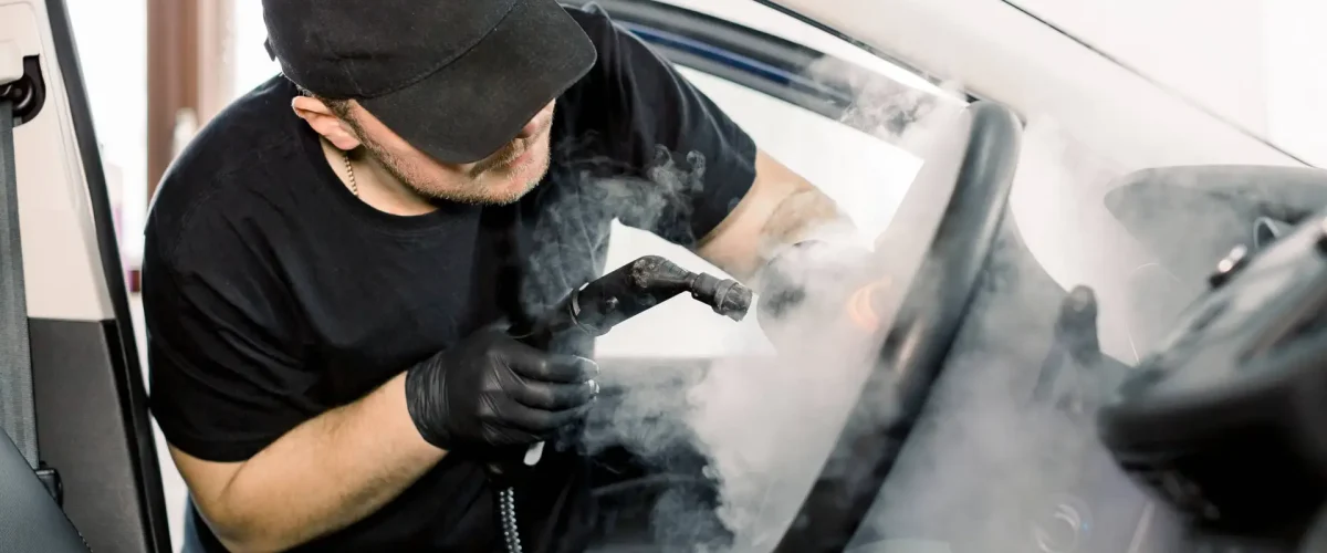 Car detailing, steam cleaning concept. Handsome man in black t-shirt and cap, worker of car wash center, cleaning car interior with hot steam cleaner. Car detailing concept.